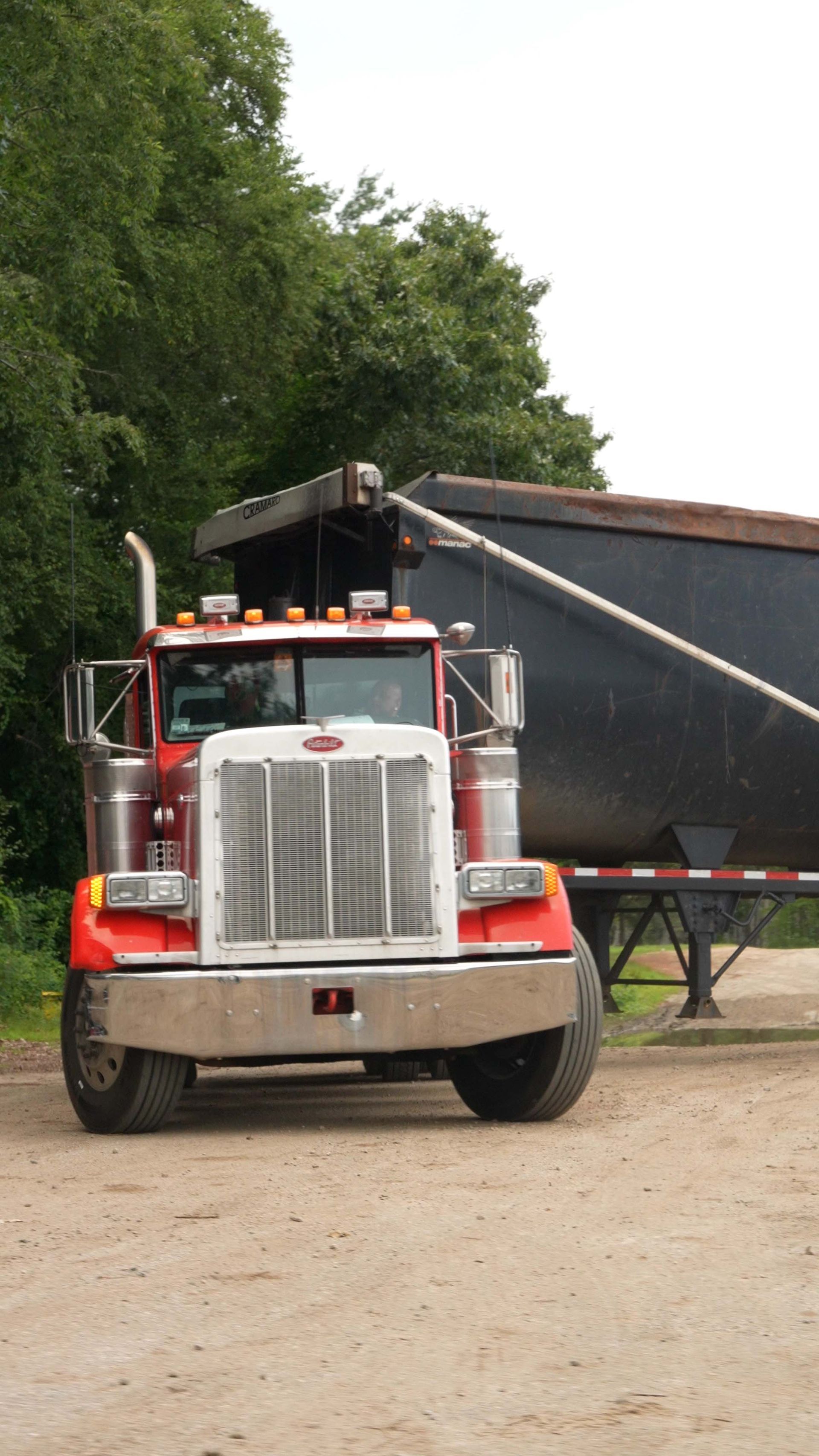 Red semi truck parked on a dirt road beside trees, hauling a large black trailer.