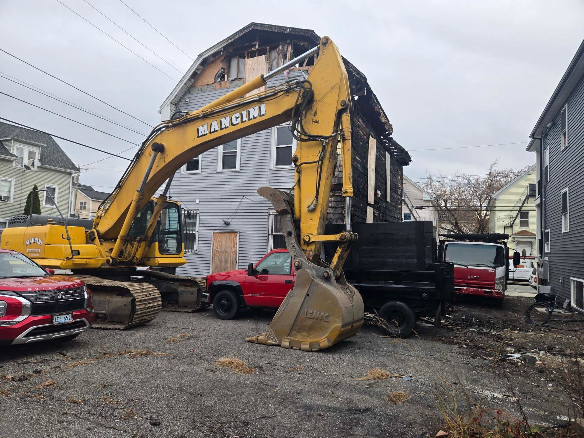 Yellow excavator tearing down a building on a residential street beside parked cars
