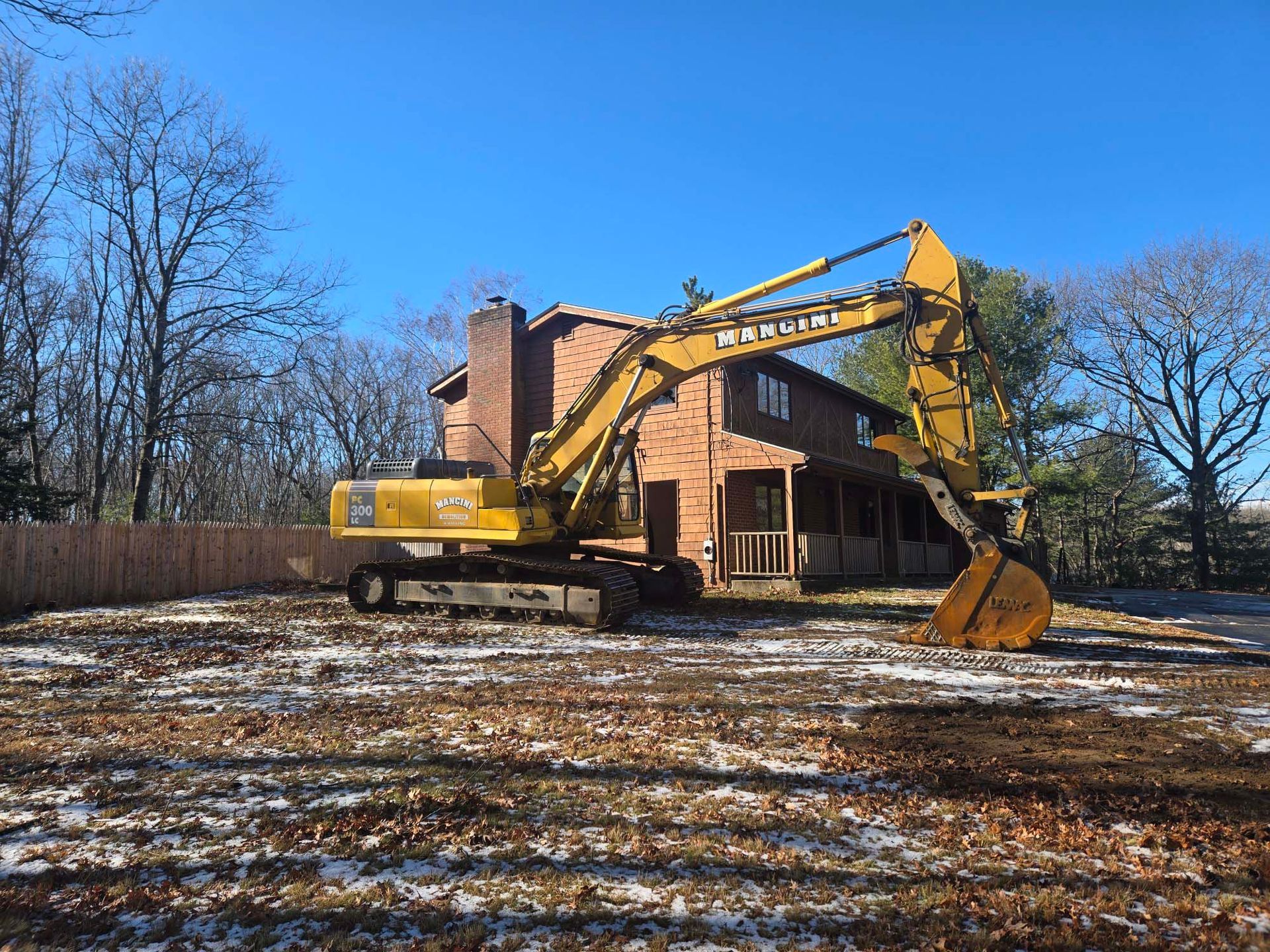 Yellow excavator demolishing a brick house on a snowy lot under a blue sky