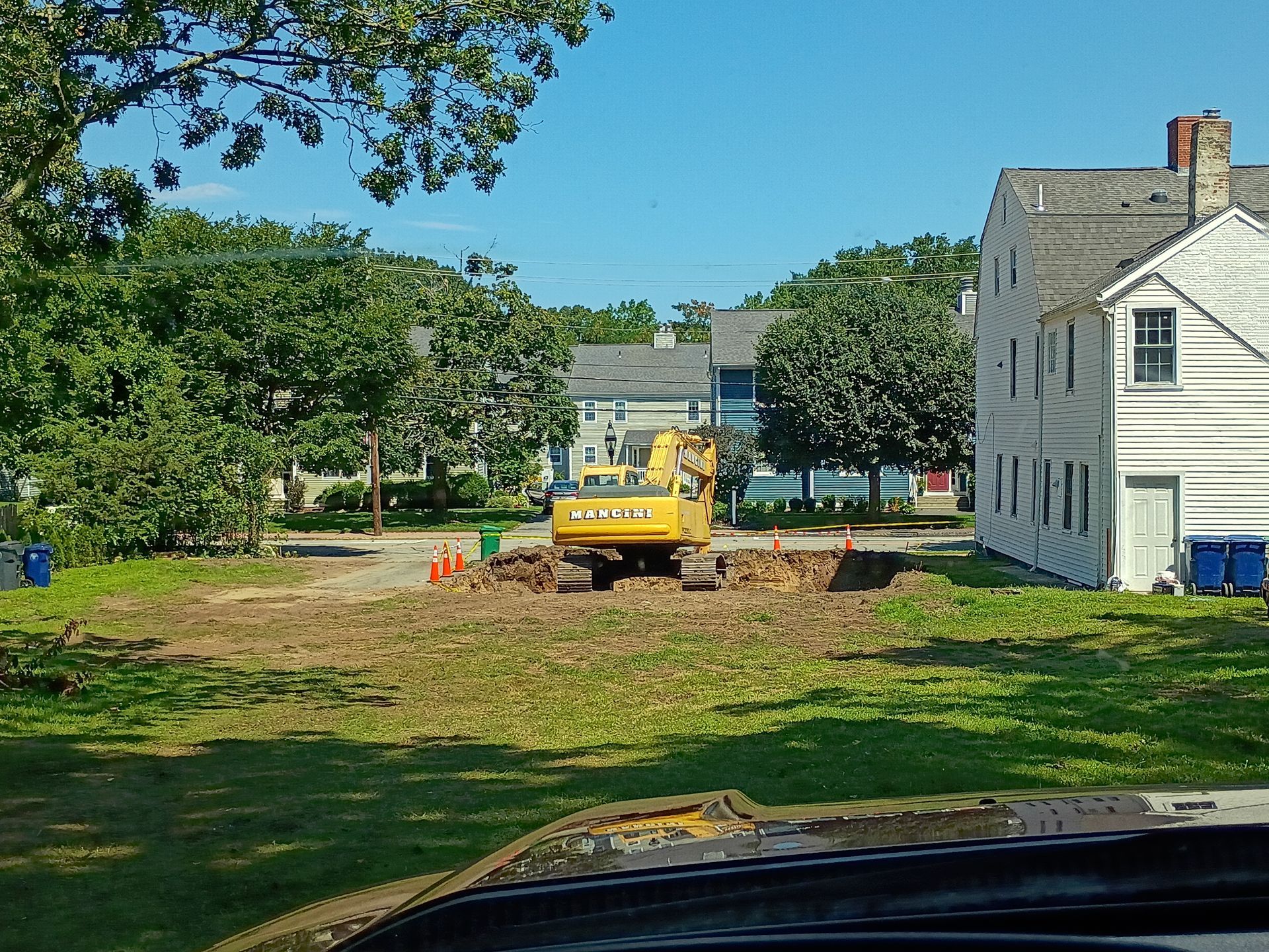 Yellow excavator clearing dirt in a grassy yard beside a white house on a sunny day