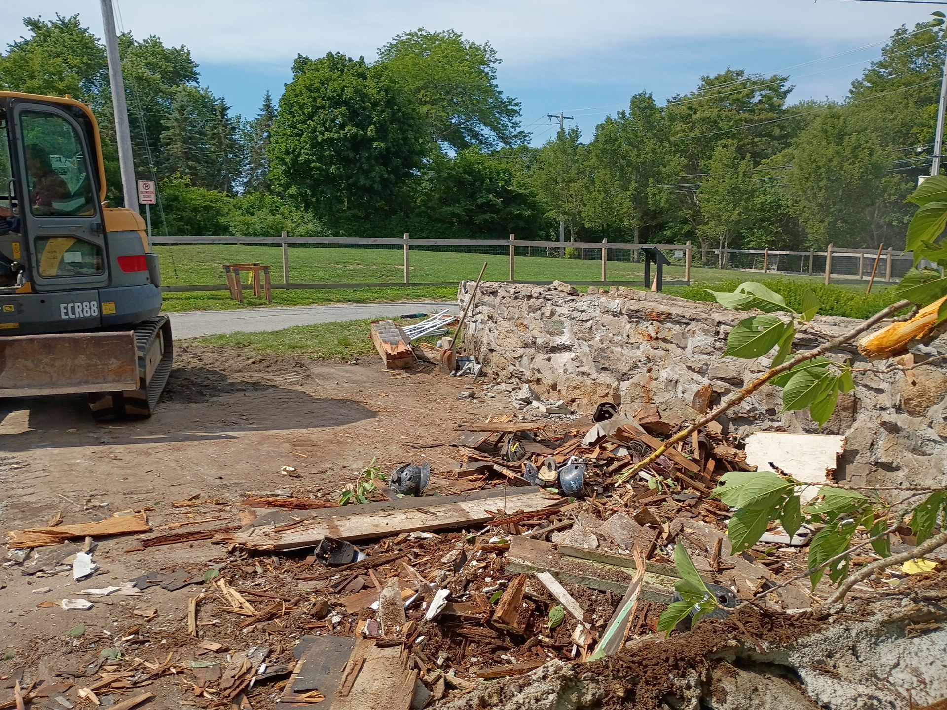 Demolition debris piled beside an excavator at a construction site with trees and fencing in the background