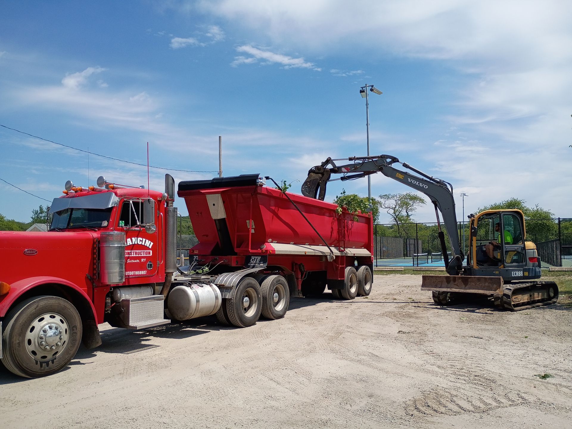 Red dump truck unloading gravel with an excavator at a construction site under a blue sky