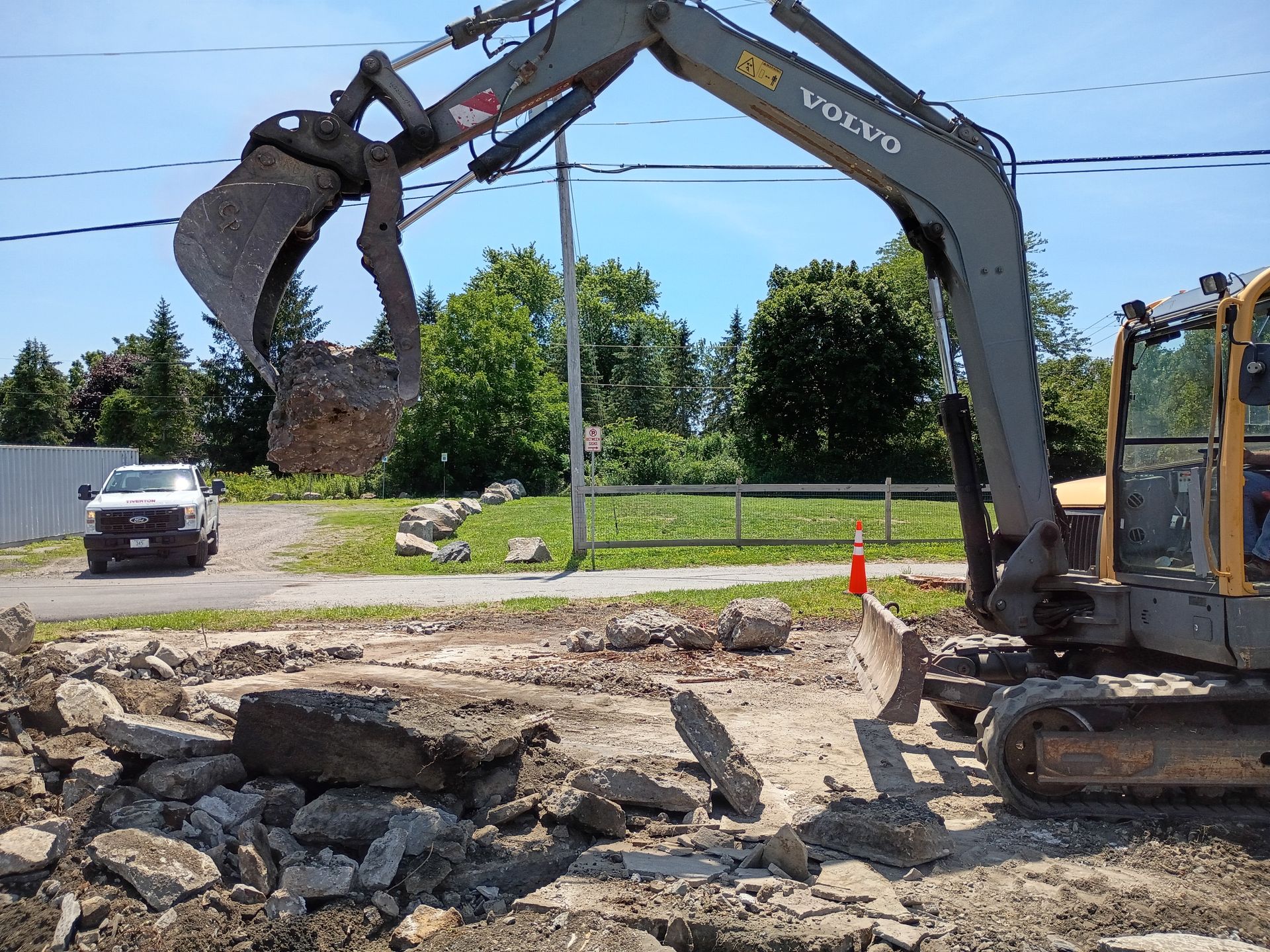 Excavator using claw to lift broken concrete on a roadside construction site with rubble and traffic cones