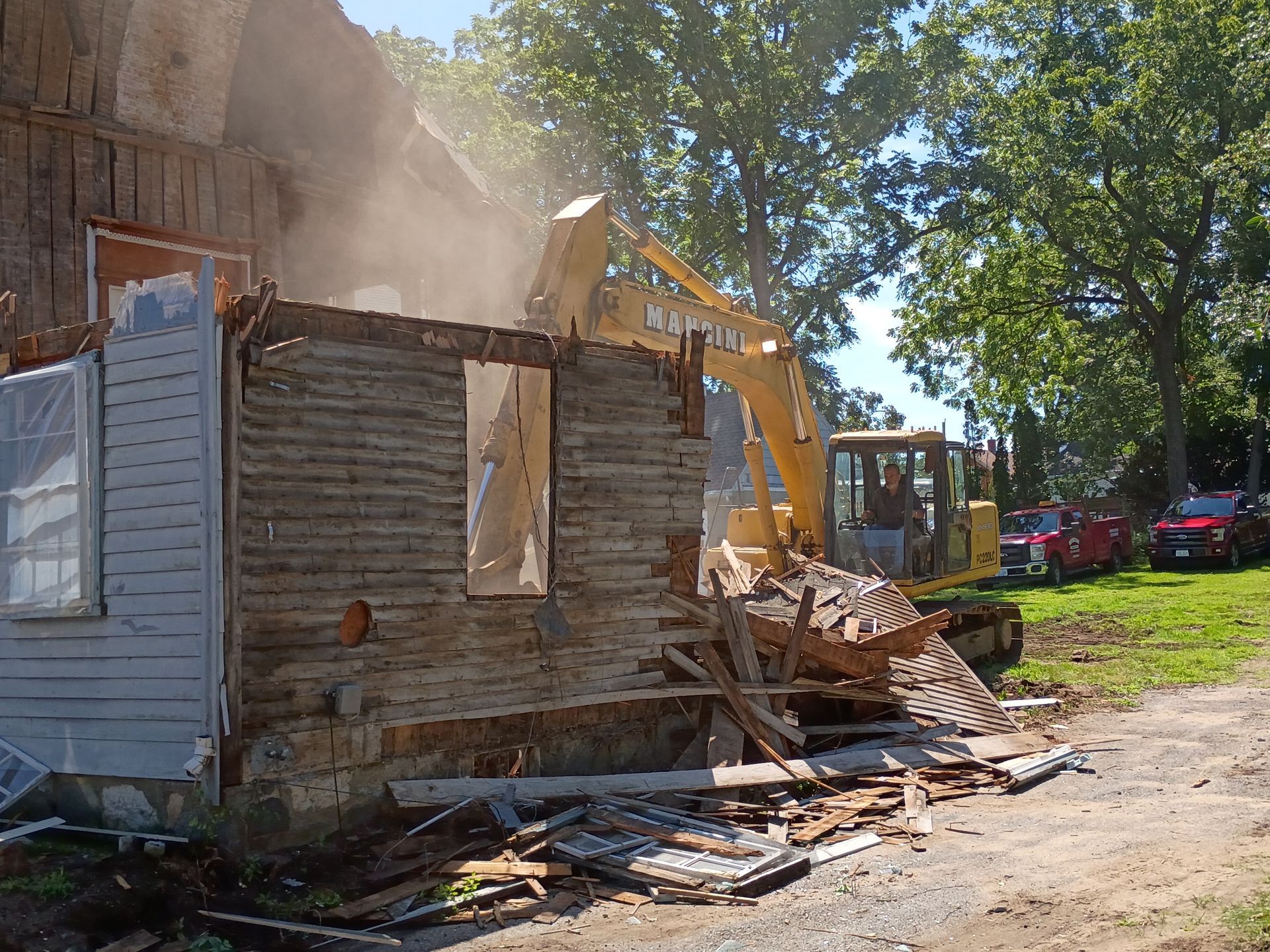 Bulldozer demolishing a small wooden house, with dust and debris in a sunny yard