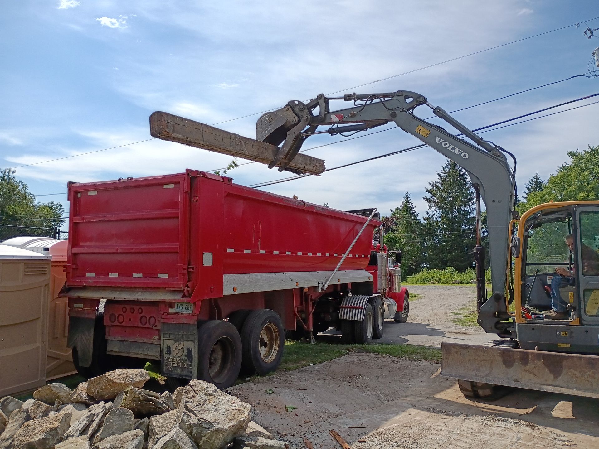 Excavator loading rocks into a red dump truck at a roadside work site