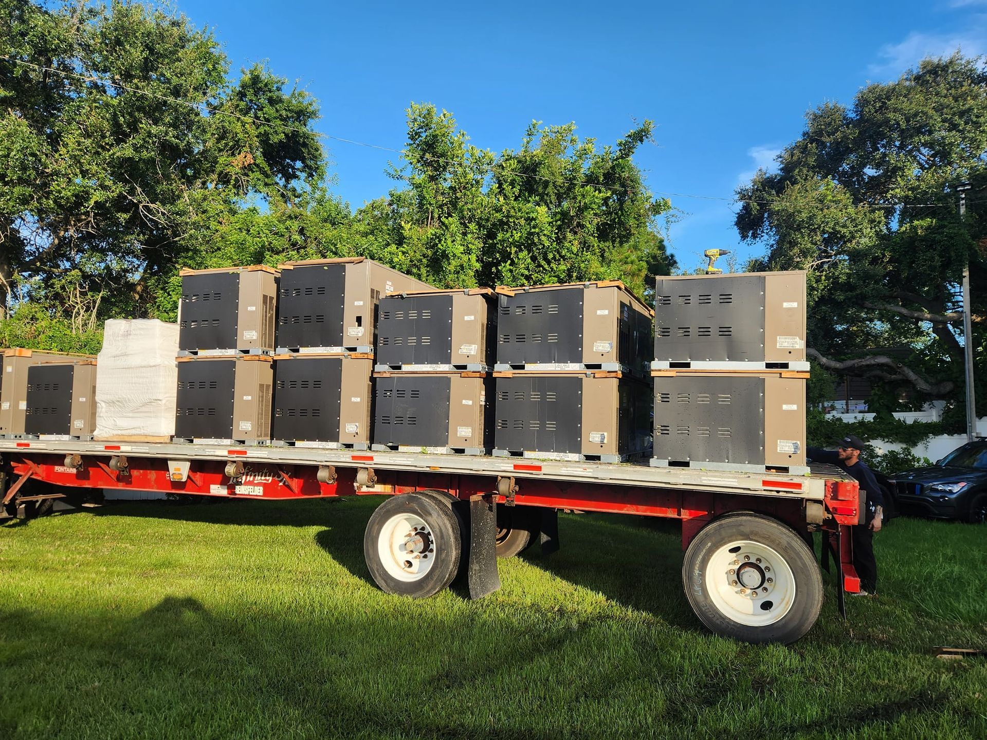 Flatbed trailer loaded with square, stacked containers on a grassy area, under a blue sky.