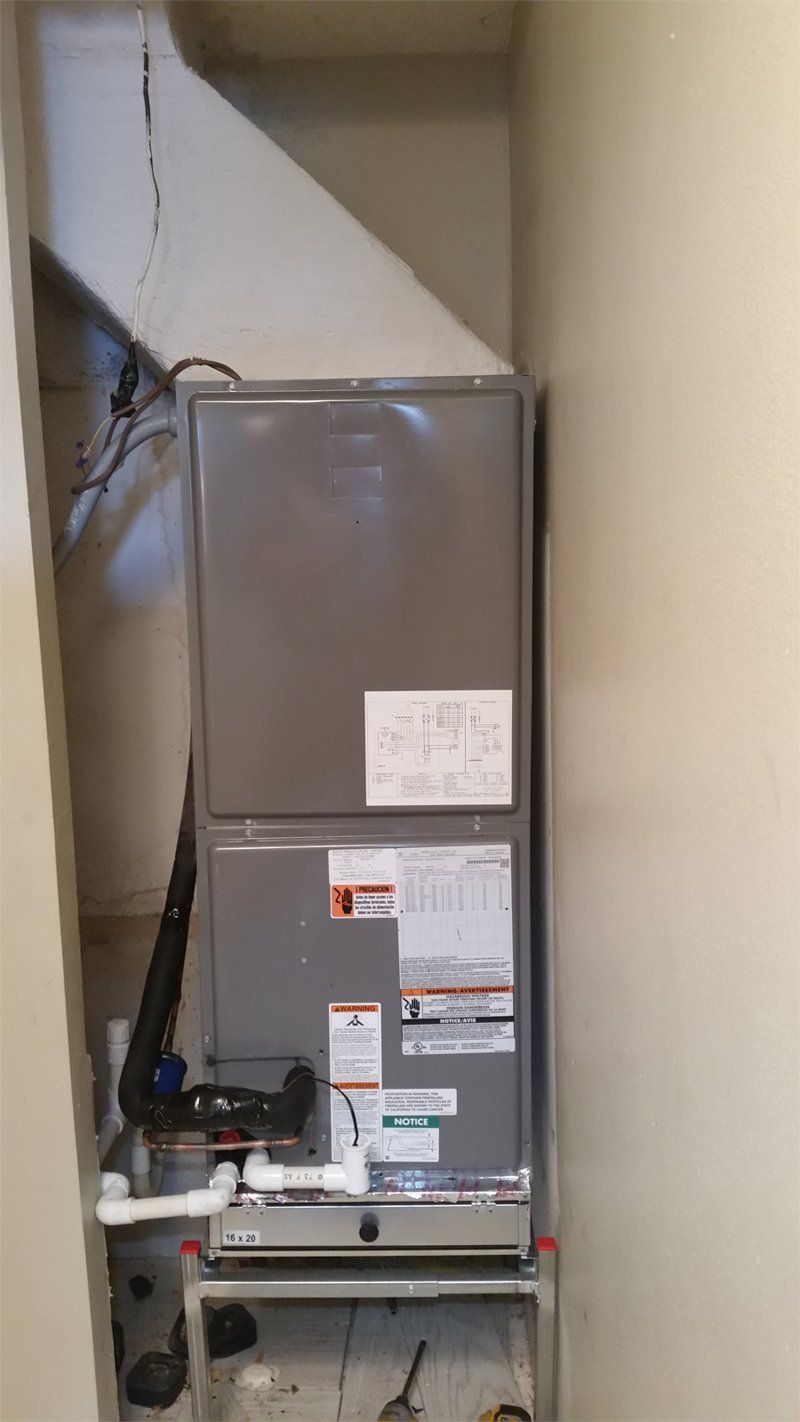 A rectangular HVAC unit, gray metal, installed in a narrow alcove. Ductwork above. Black pipe and white PVC.