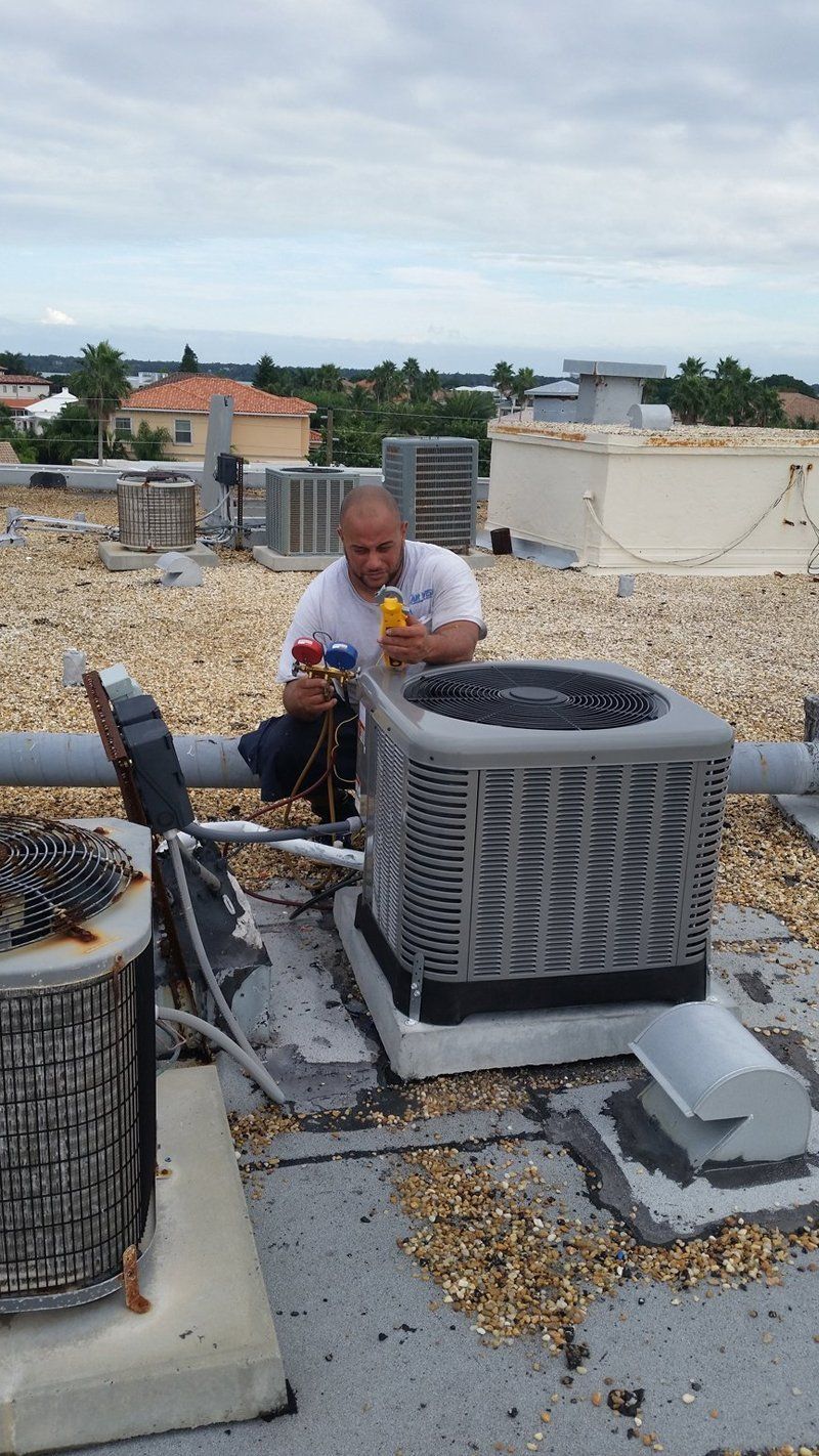 Man on rooftop servicing an air conditioning unit. He's holding gauges. Other units and a cityscape are in the background.