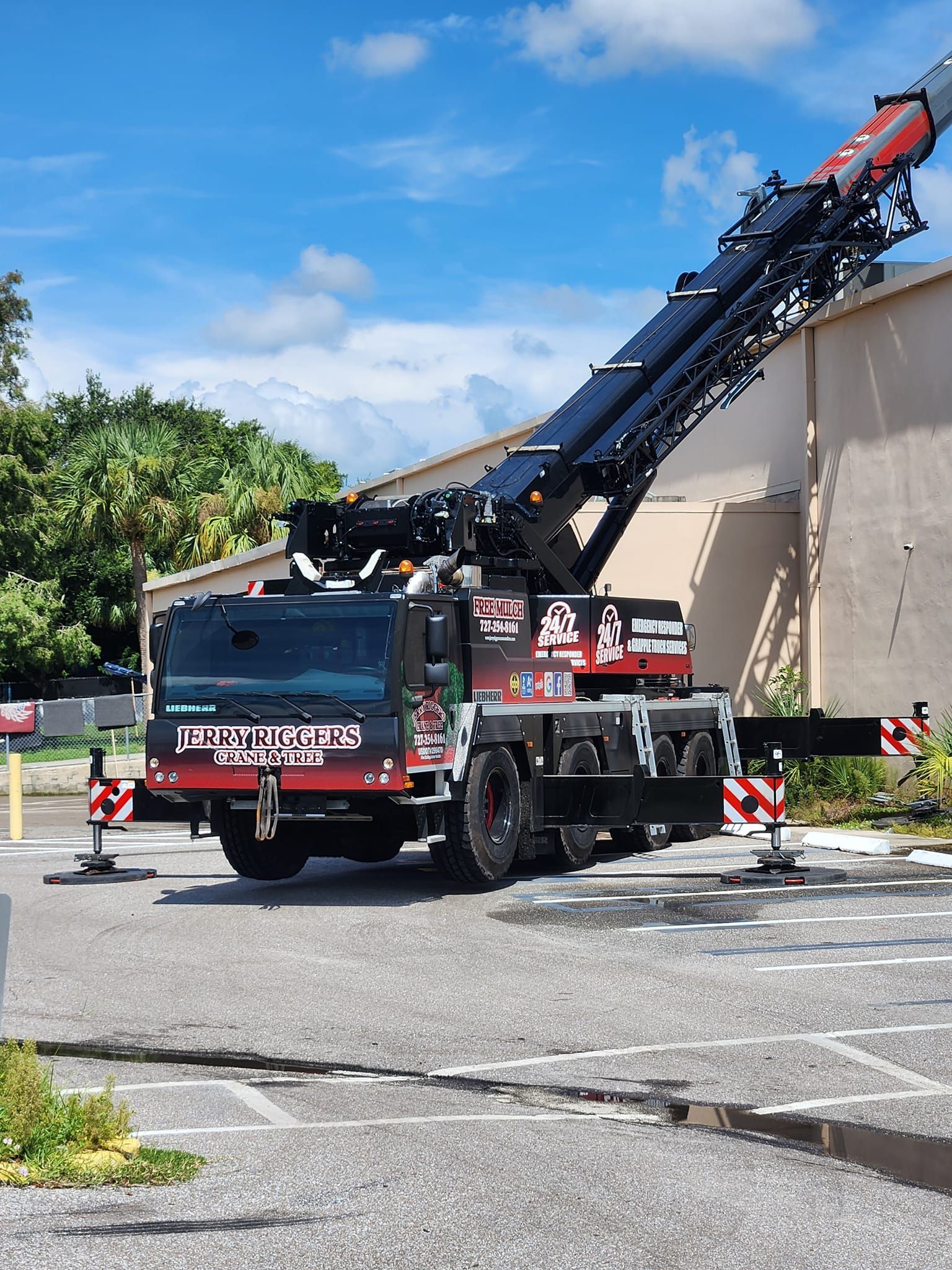Large red and black crane vehicle with its arm extended, parked on asphalt in front of a building under a blue sky.