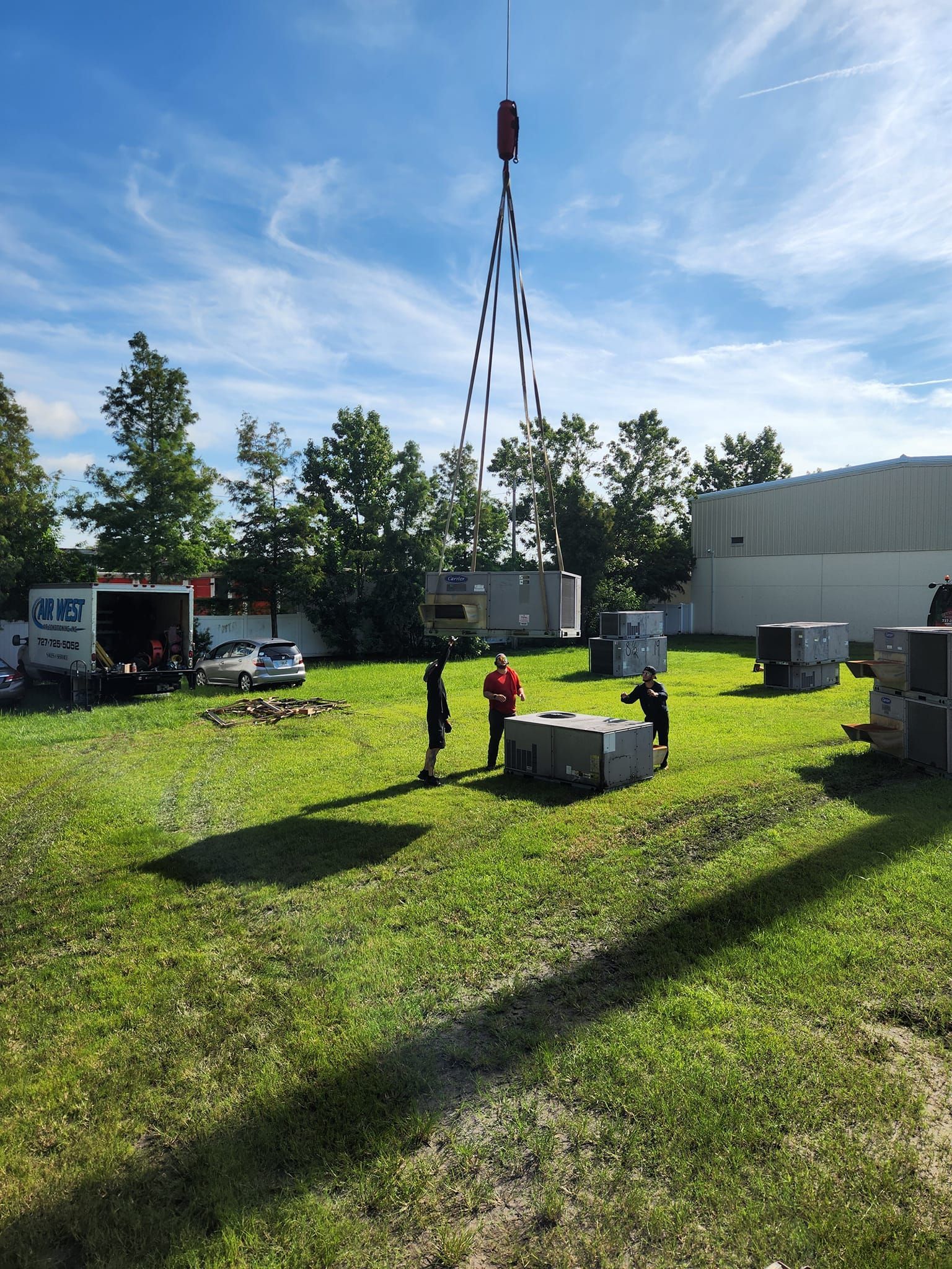 Crane lifting a large gray box on a grassy field; workers supervise. White building, trees, and blue sky in the background.