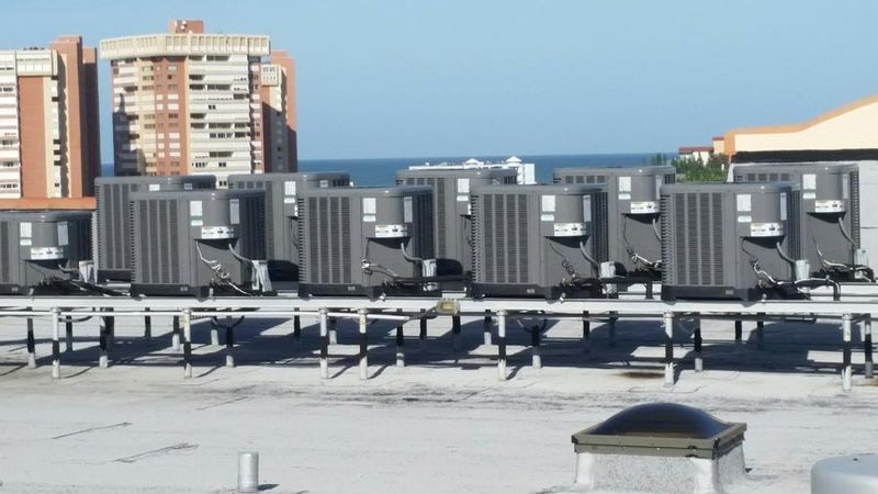 Row of air conditioning units on a rooftop, with a building and ocean in the background.