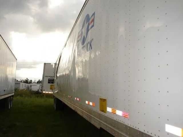 White semi-trailer with blue and white logo, parked on grass under a cloudy sky.