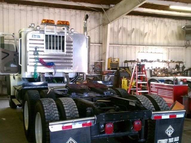 Rear view of a semi-truck cab in a garage. Silver and black, with safety lights and a work area in background.
