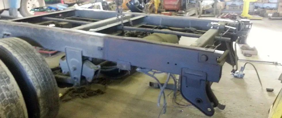 Truck chassis in a workshop setting, tires visible, showing frame and suspension components.