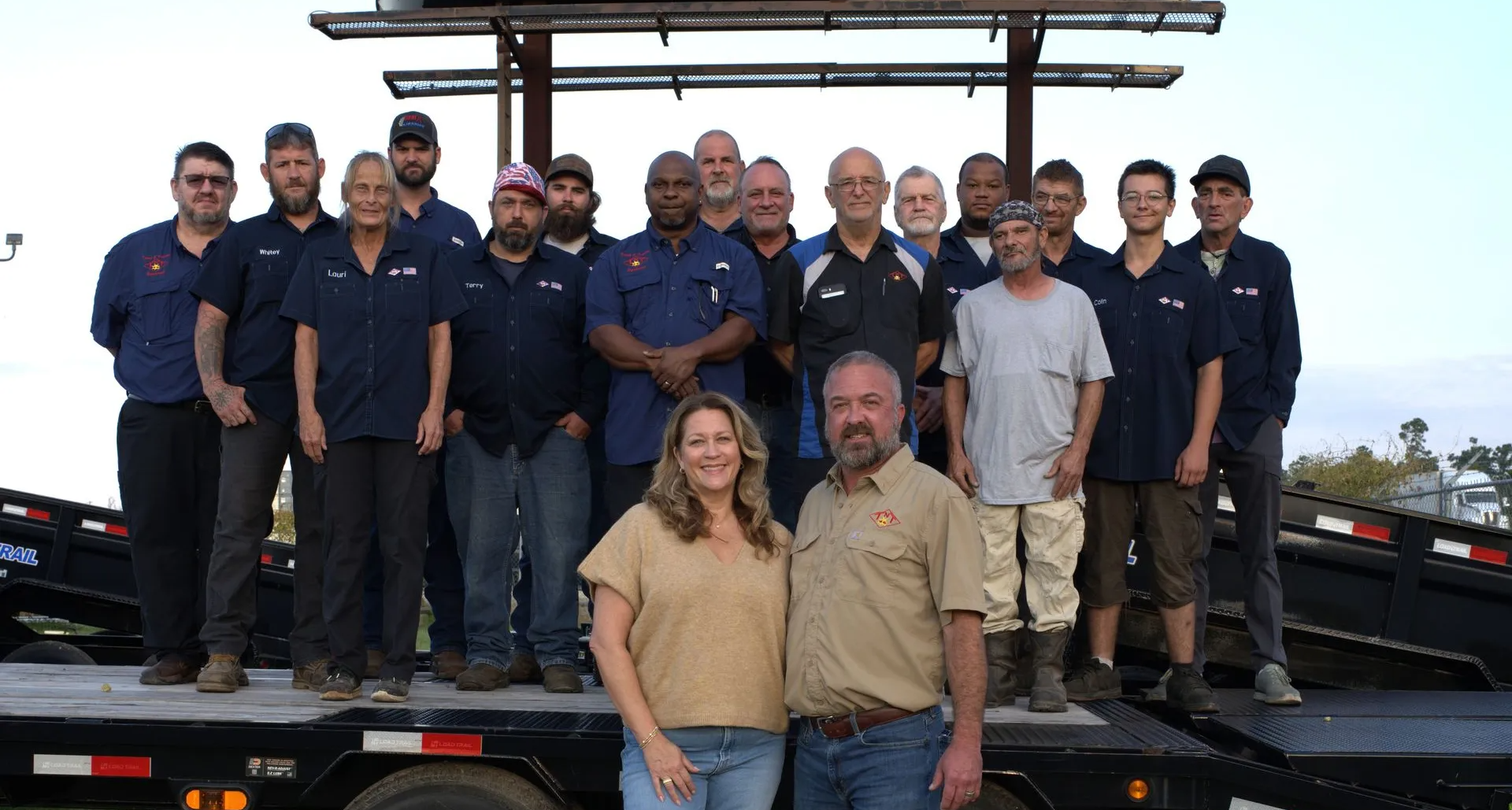 Group of people, including two in front, posing on a trailer. Many wear work attire, under a metal structure.