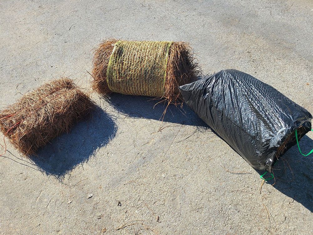 Three bundles of pine straw on concrete; one wrapped in netting, one in a black bag, and one loose.