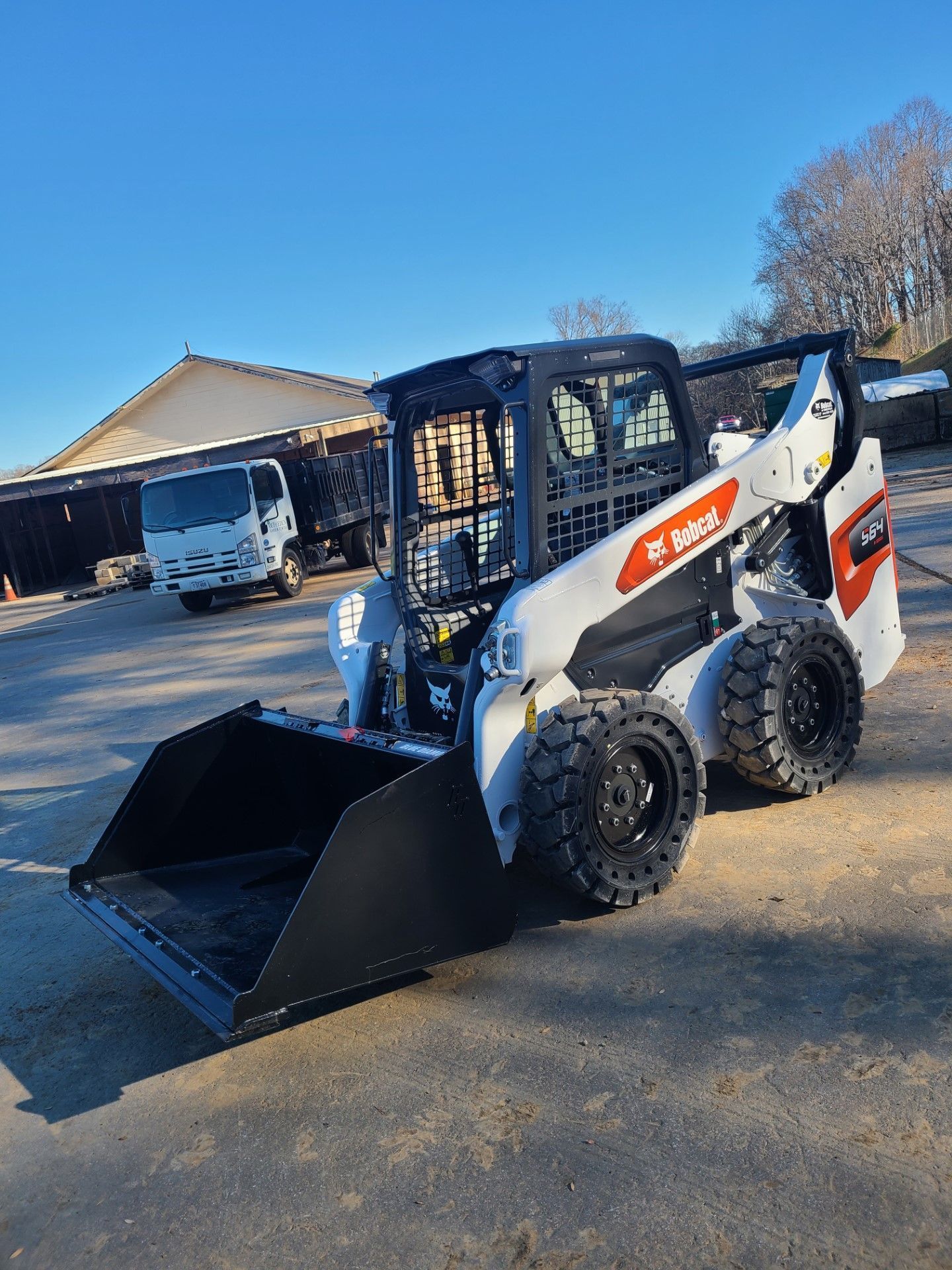 Bobcat skid-steer loader with a black bucket, parked outdoors on asphalt, against a backdrop of a building and truck.