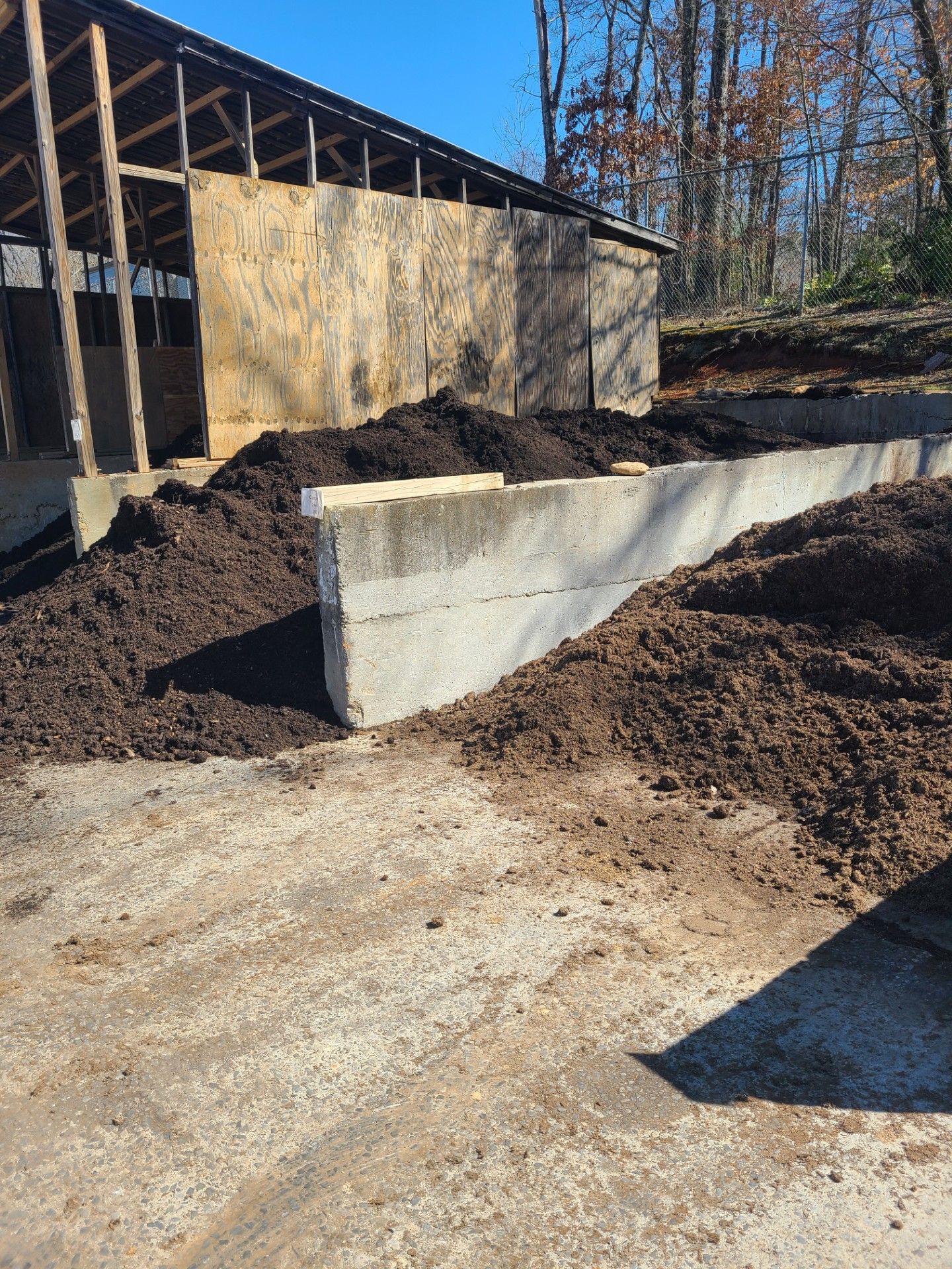 Concrete retaining wall with large piles of dark mulch in front of a partially constructed wooden building.