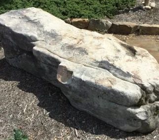 Large, light gray stone bench in a garden setting, with shadows and greenery.
