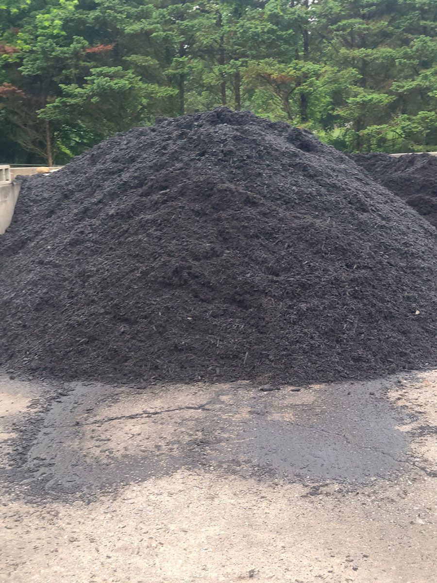 Large pile of black mulch on a concrete surface, with trees in the background.