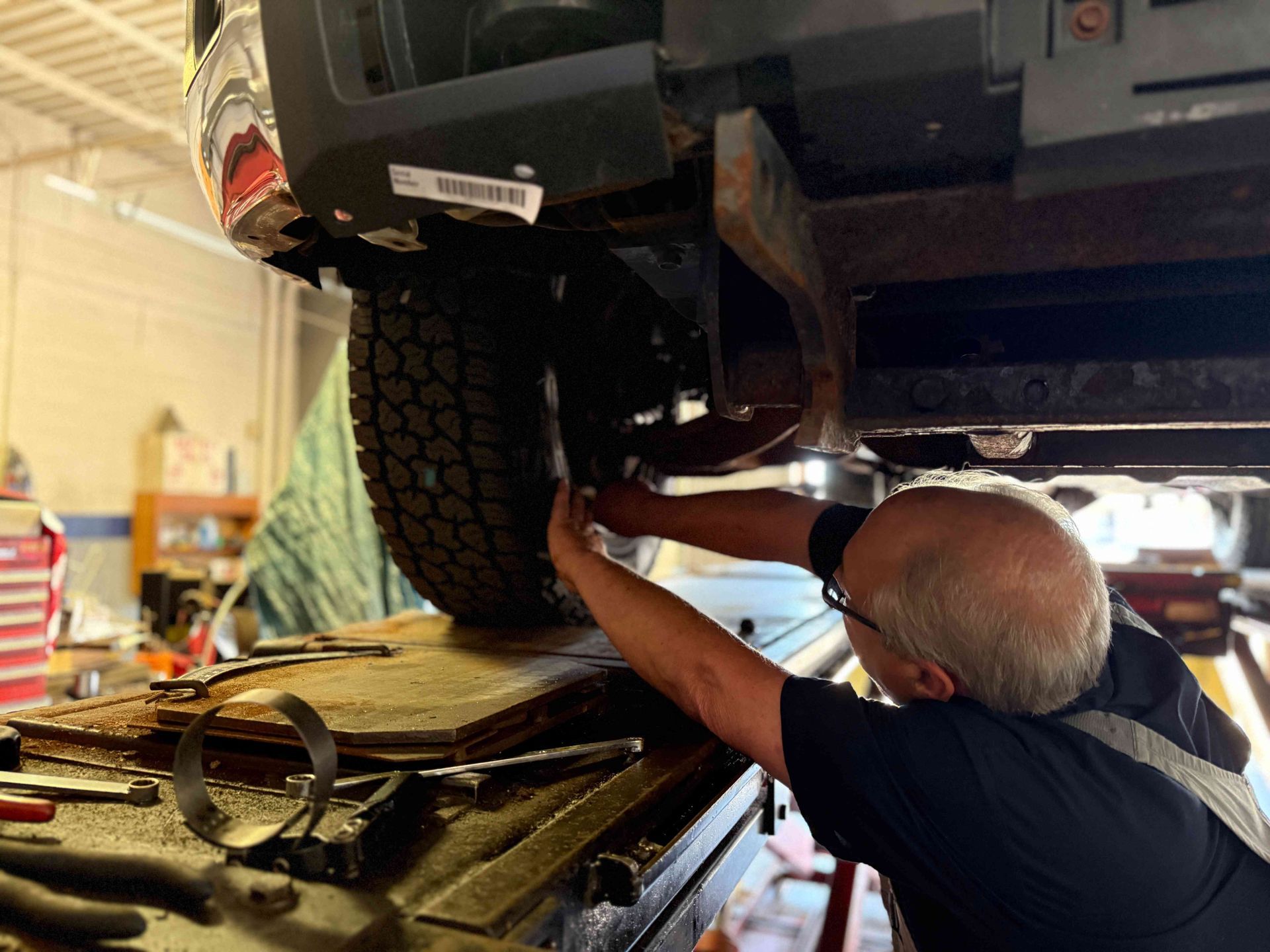 Mechanic working under a lifted car in a garage, using tools near the rear wheel.