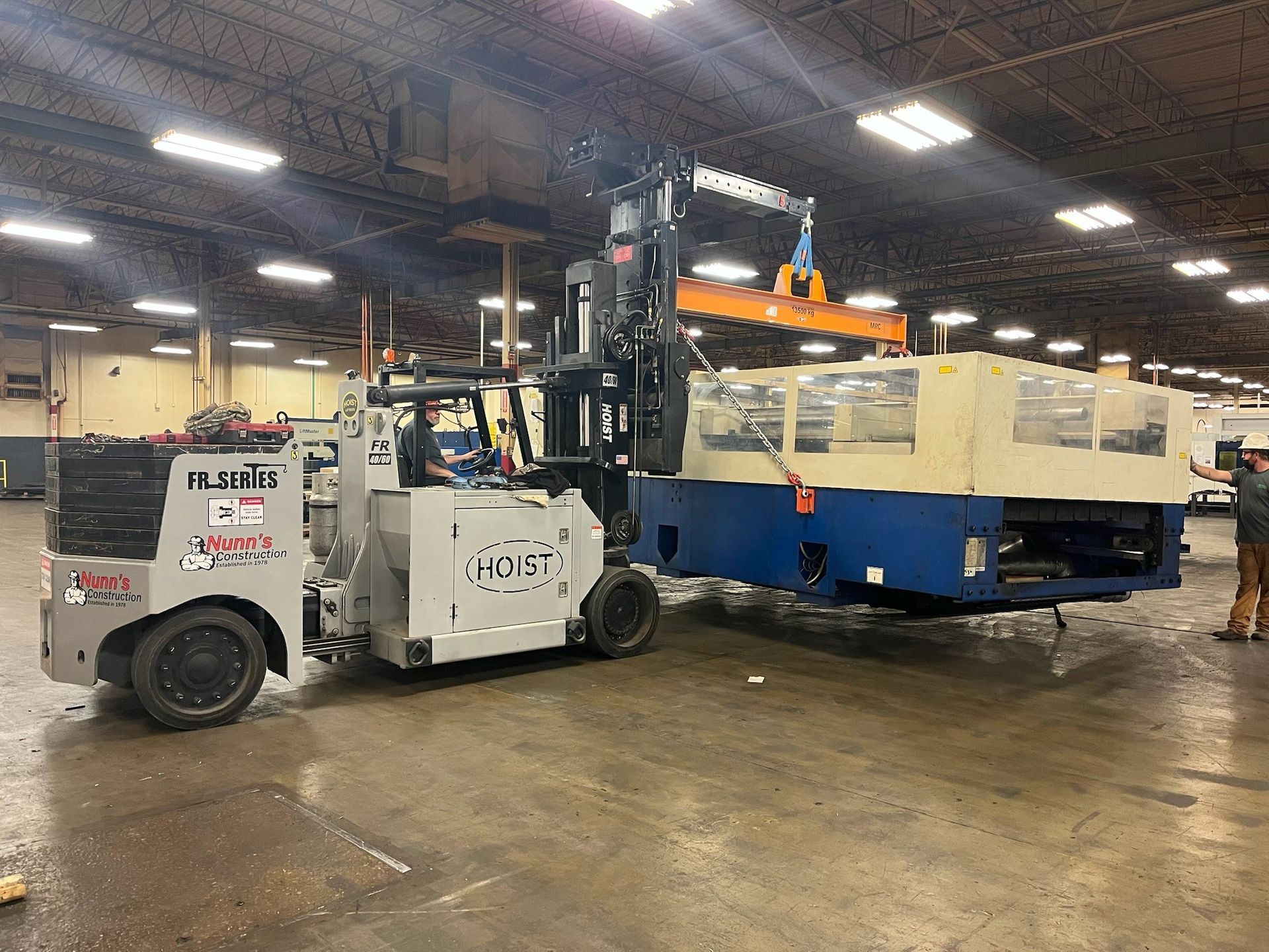 Forklift moving industrial equipment in a warehouse, another lift assists.