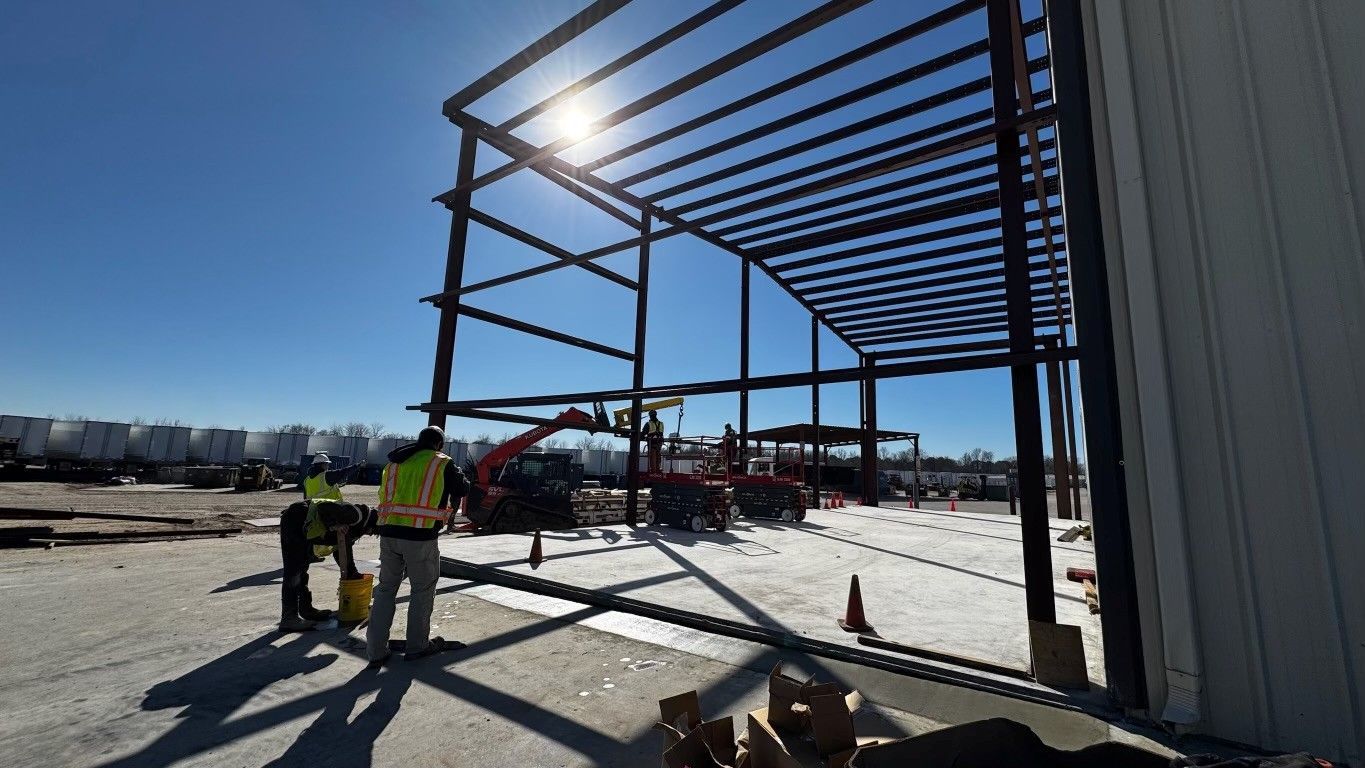 Construction of a steel-framed building on a sunny day. Workers and machinery visible. Blue sky.