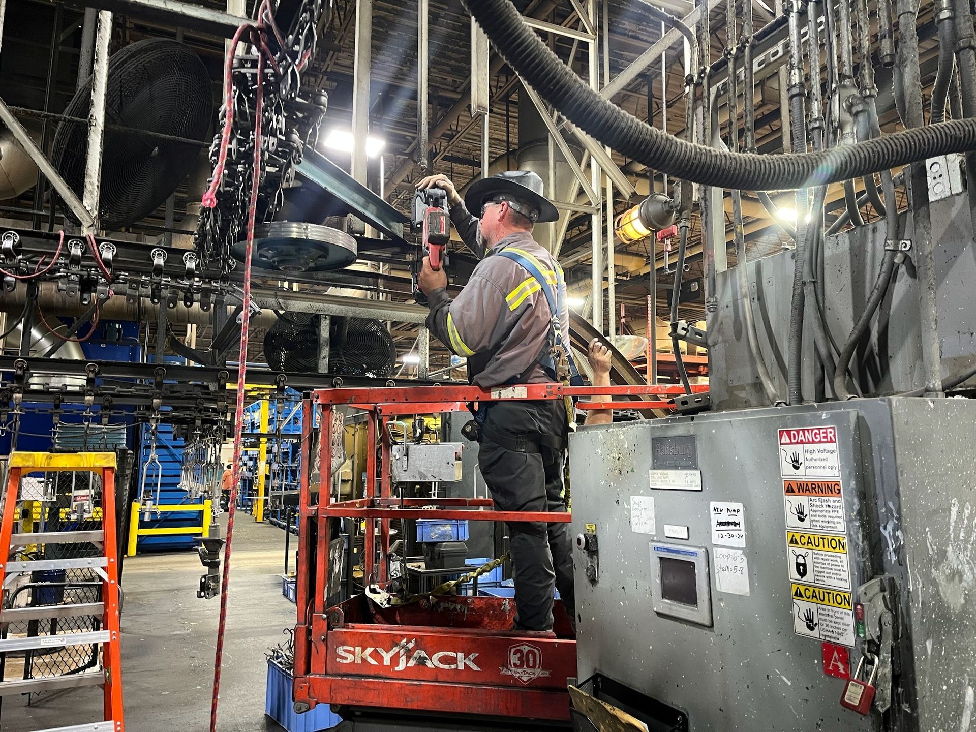 Worker on lift in factory; repairs equipment. Wears hard hat, safety vest. Industrial setting, machinery visible.