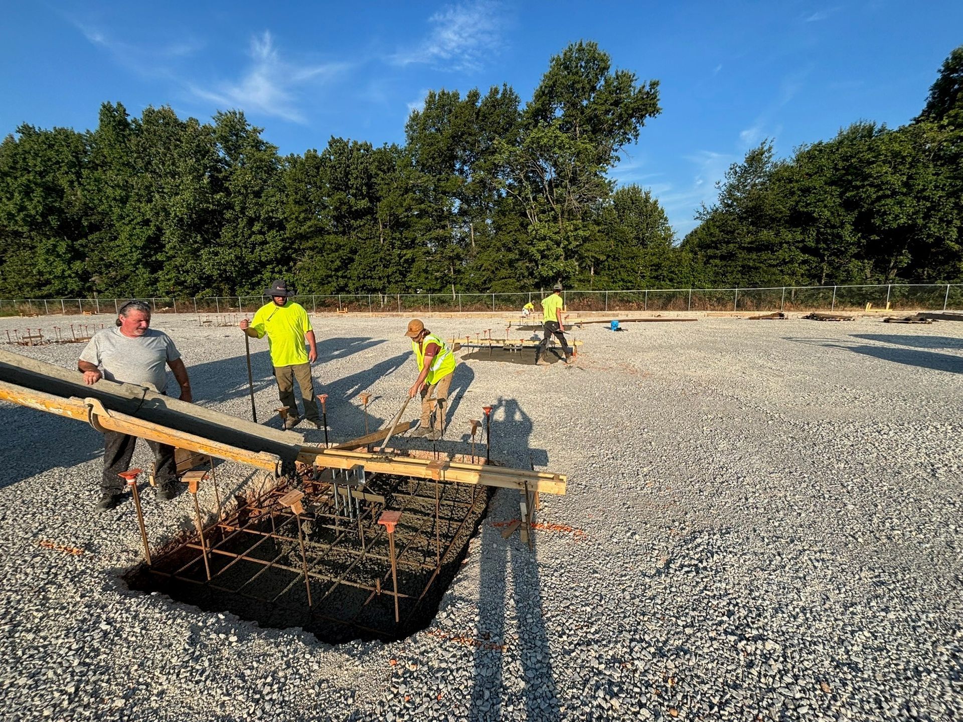 Construction workers leveling concrete on a gravel surface. They are wearing safety vests and surrounded by trees.