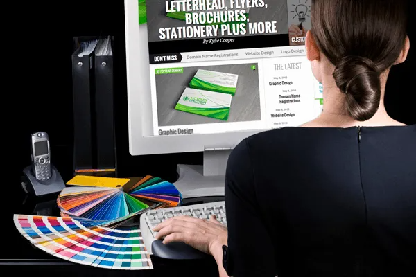 Person viewing a desktop monitor with colorful print samples and a brochure design on screen