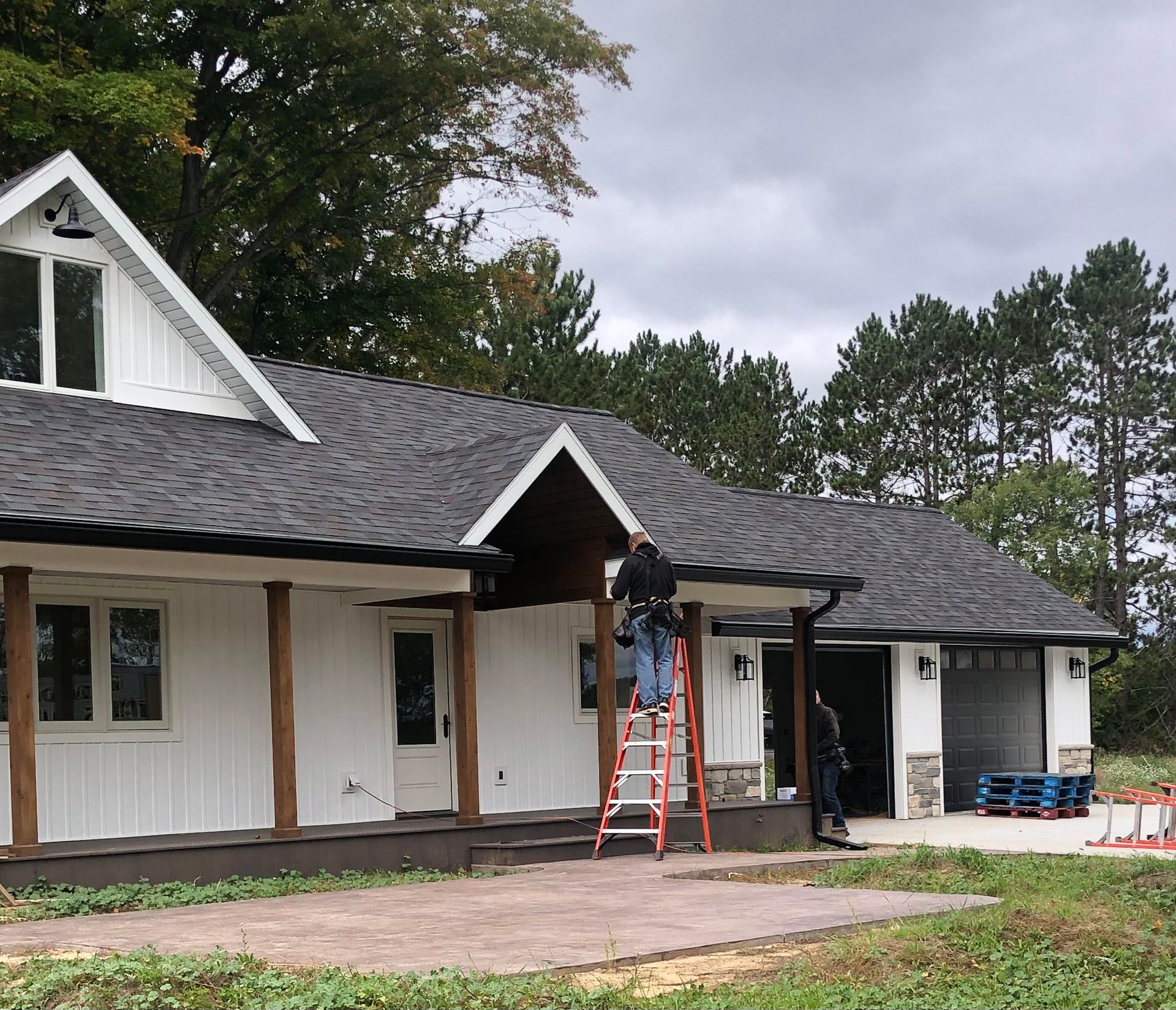 Workers on a ladder installing gutters on a white house with a dark roof and garage. Cloudy day.