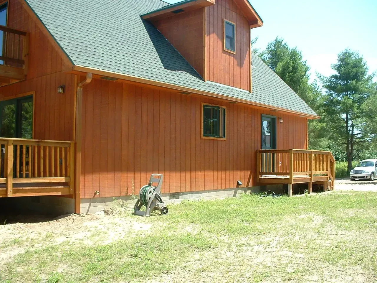 Brown wooden house with green roof and deck in a grassy yard on a sunny day.