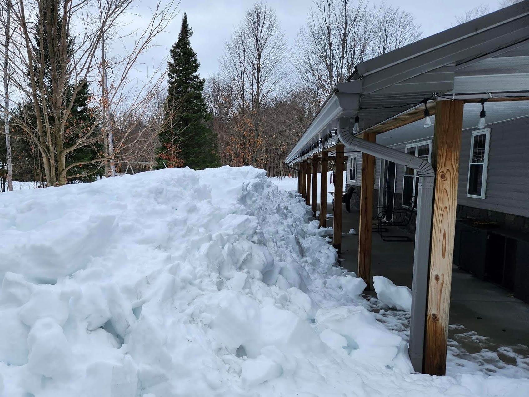 Snow piled high next to a house with a porch. Overcast day.