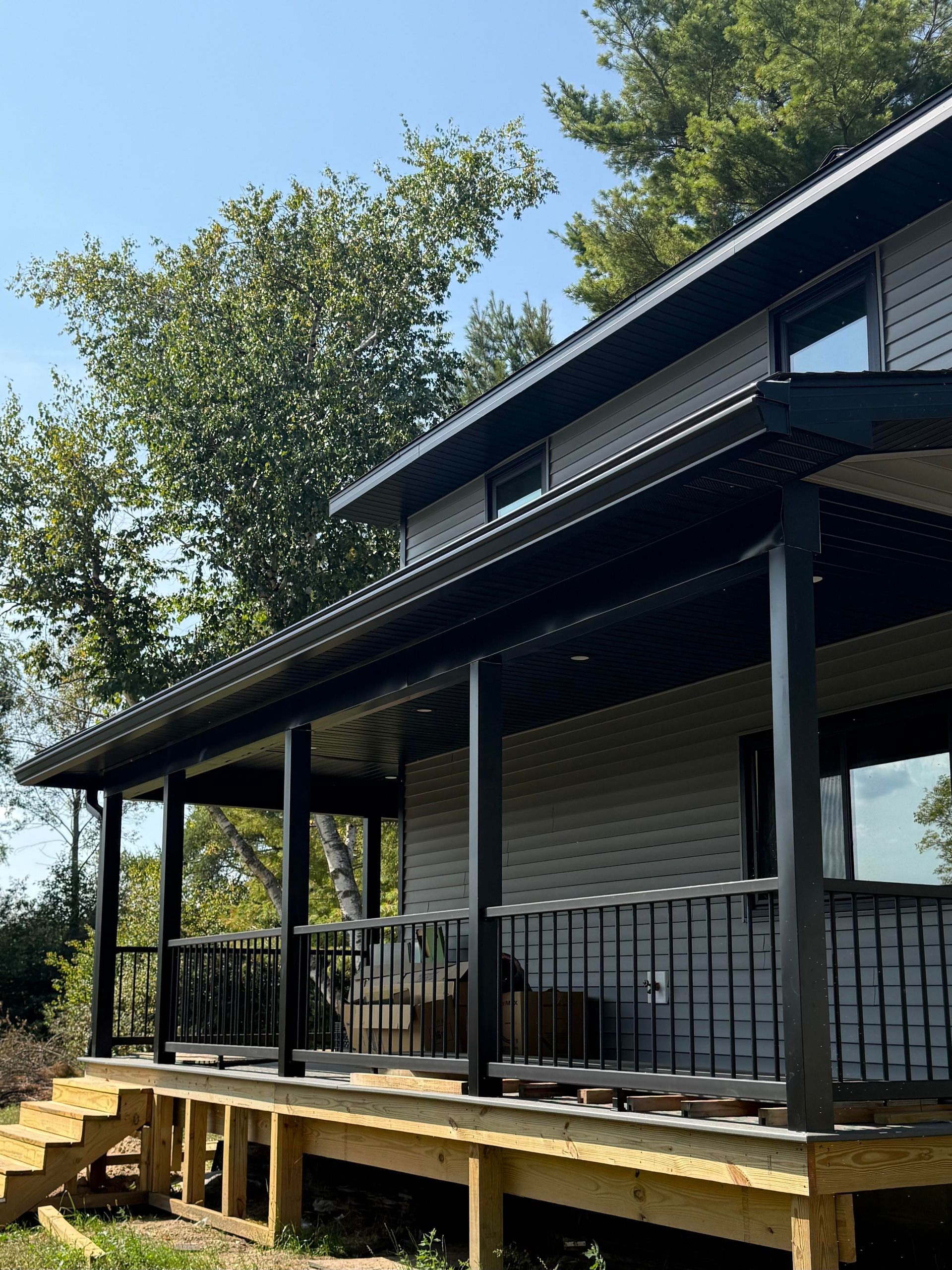Two-story house with a black-trimmed porch and gray siding, wooden deck, stairs, and foliage.