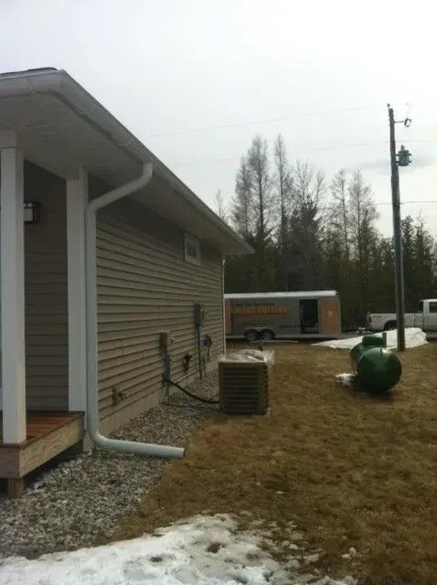 Beige building with white trim, gutter, and AC unit; trailer, trees, and snow on the ground in the background.