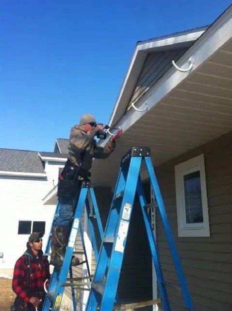 Two men installing gutters on a house, one on a ladder using a power tool, the other watching.