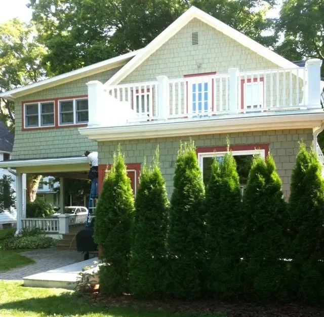 Green house with white railing, red trim, and line of green trees in front.