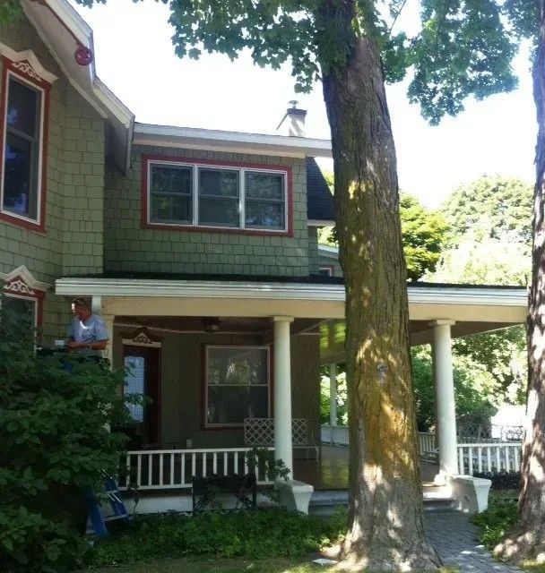 Two-story green house with porch, white columns. Person on porch, trees in front.