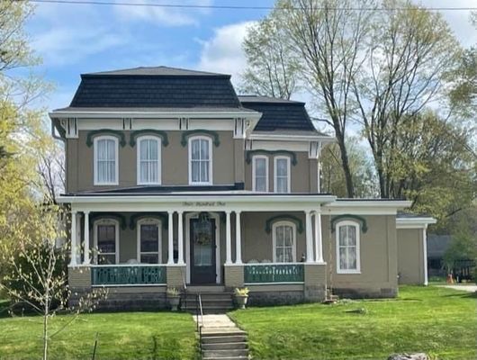 Two-story tan house with black roof, white trim, and green porch, set on green lawn with trees.