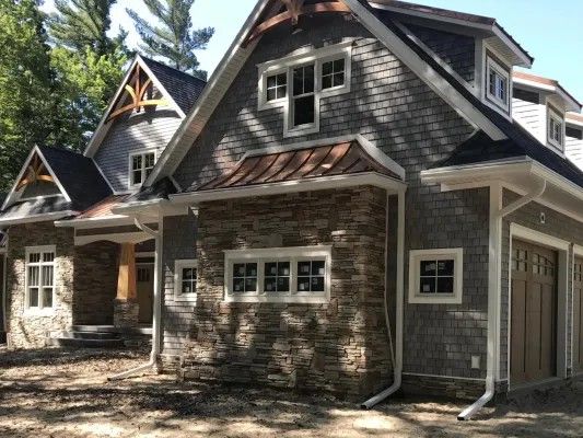 Gray and brown craftsman-style house with stone accents, shake siding, and copper details, set in a wooded area.