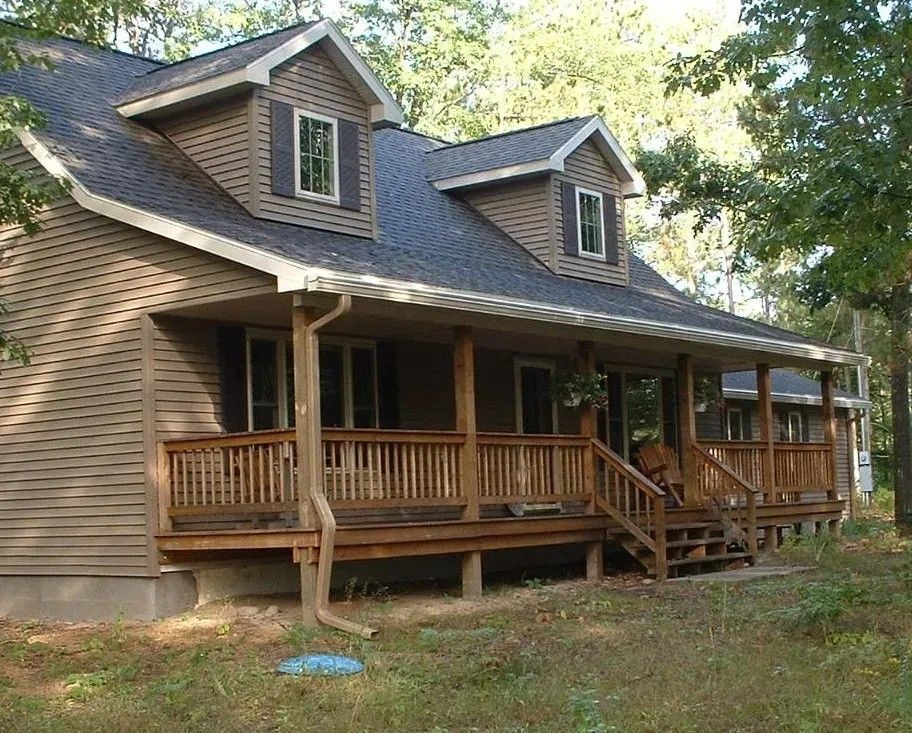 Beige house with a wraparound wooden porch and dark gray roof in a wooded area.