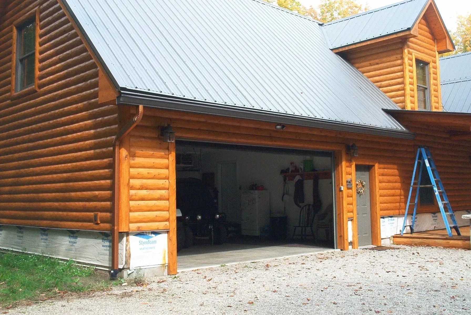 Log cabin garage with open door, gray roof, and blue ladder.