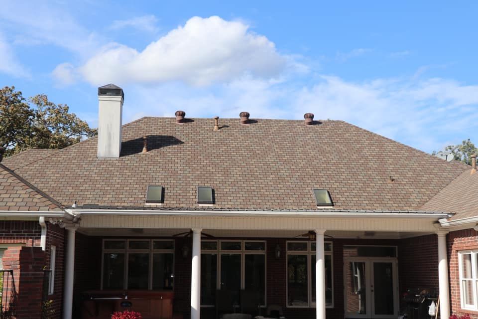 Brown shingled roof with a chimney and vents, above a brick building with a porch and windows against a blue sky.