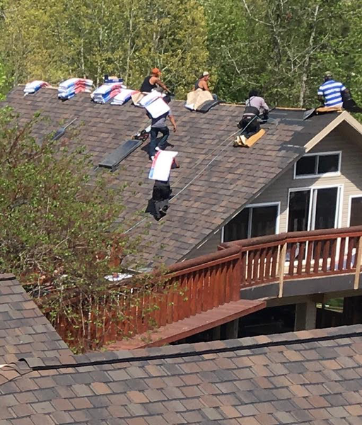 Roofers working on a house roof, carrying materials. Brown roof shingles, wooden deck and trees in the background.