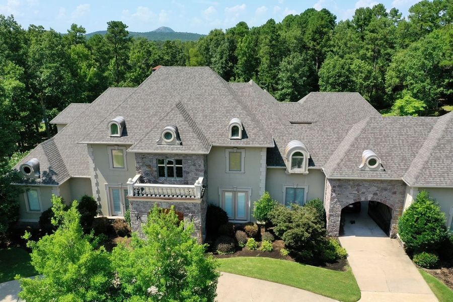 Large gray-roofed house with stone accents, balcony, and driveway, surrounded by trees.
