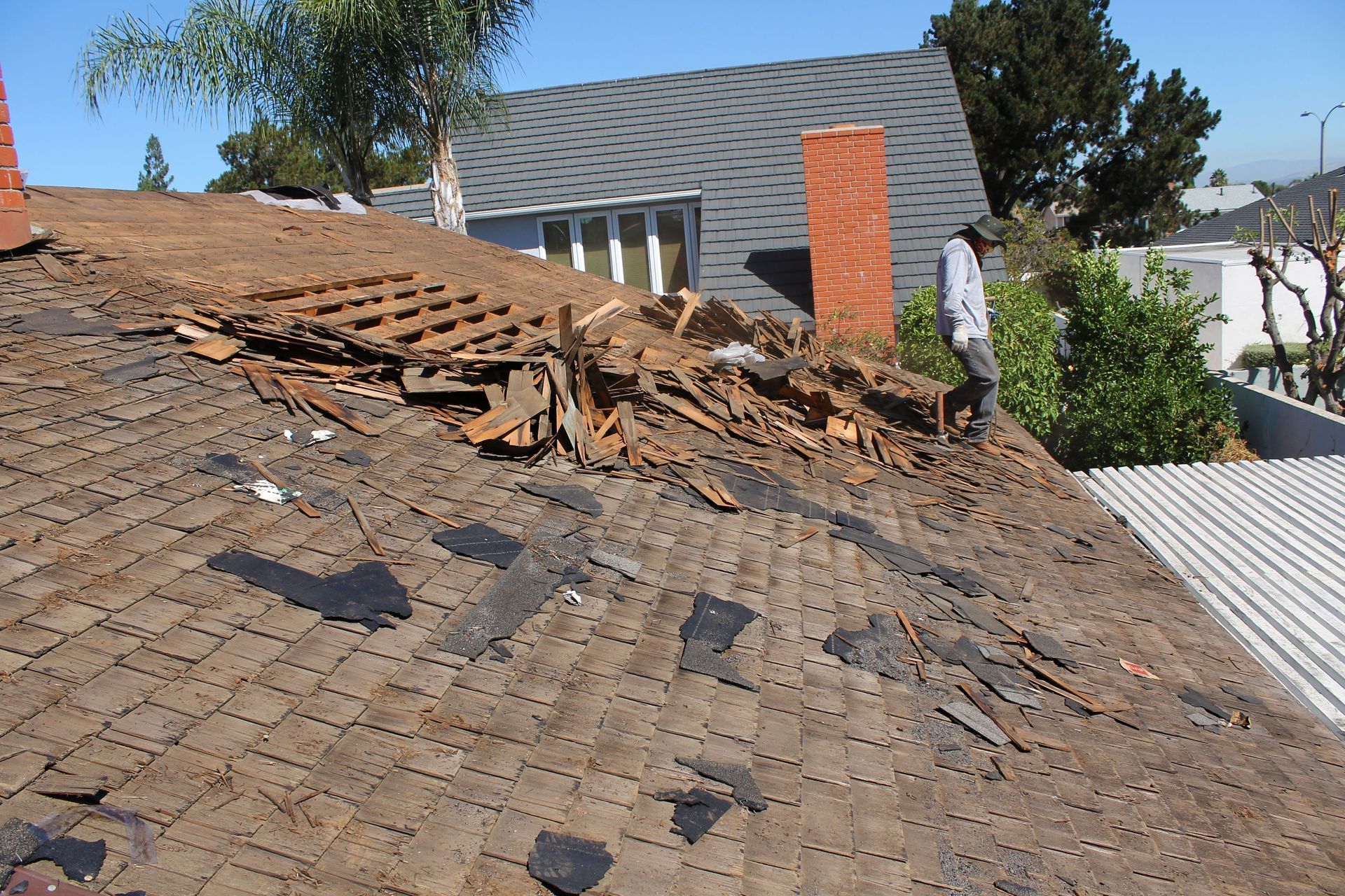 Damaged roof with exposed underlayment; person standing near chimney; sunny outdoor setting.