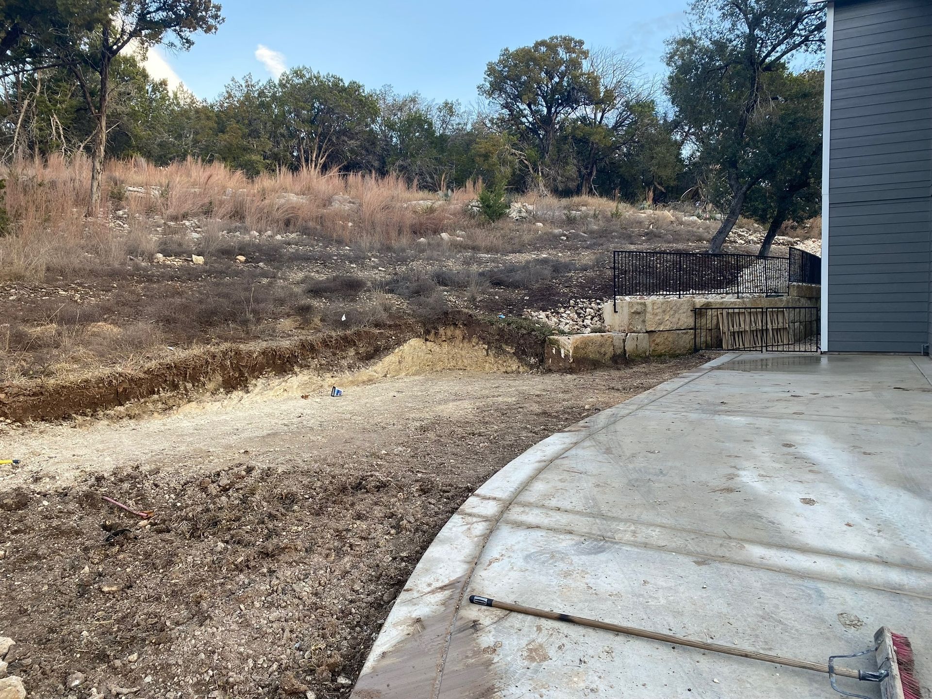 A concrete patio sits beside a yard with a low stone retaining wall and a sloped, natural hillside with trees.