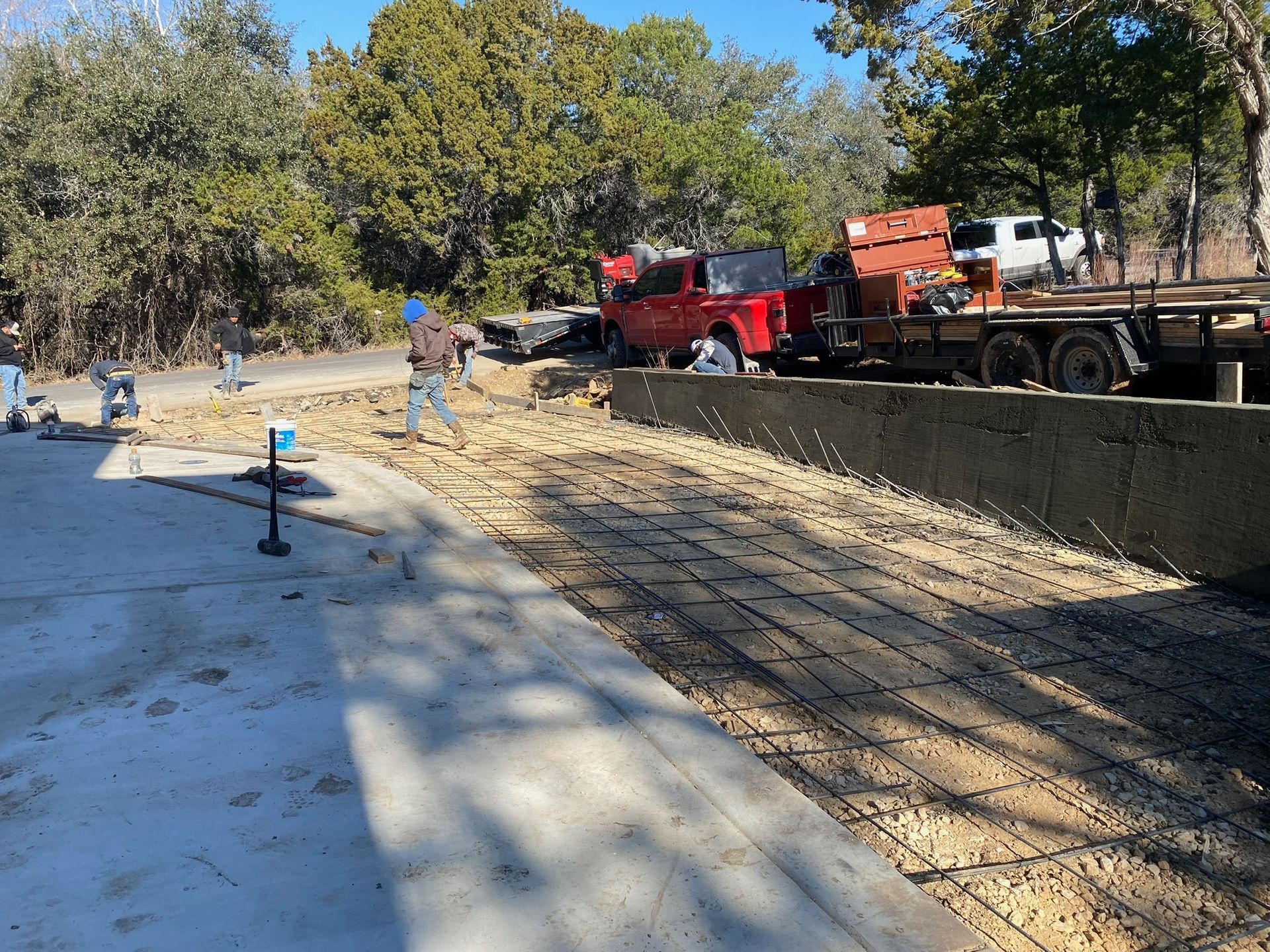 Construction workers prepare a gravel area next to a paved concrete slab, with a red truck and trailer nearby.