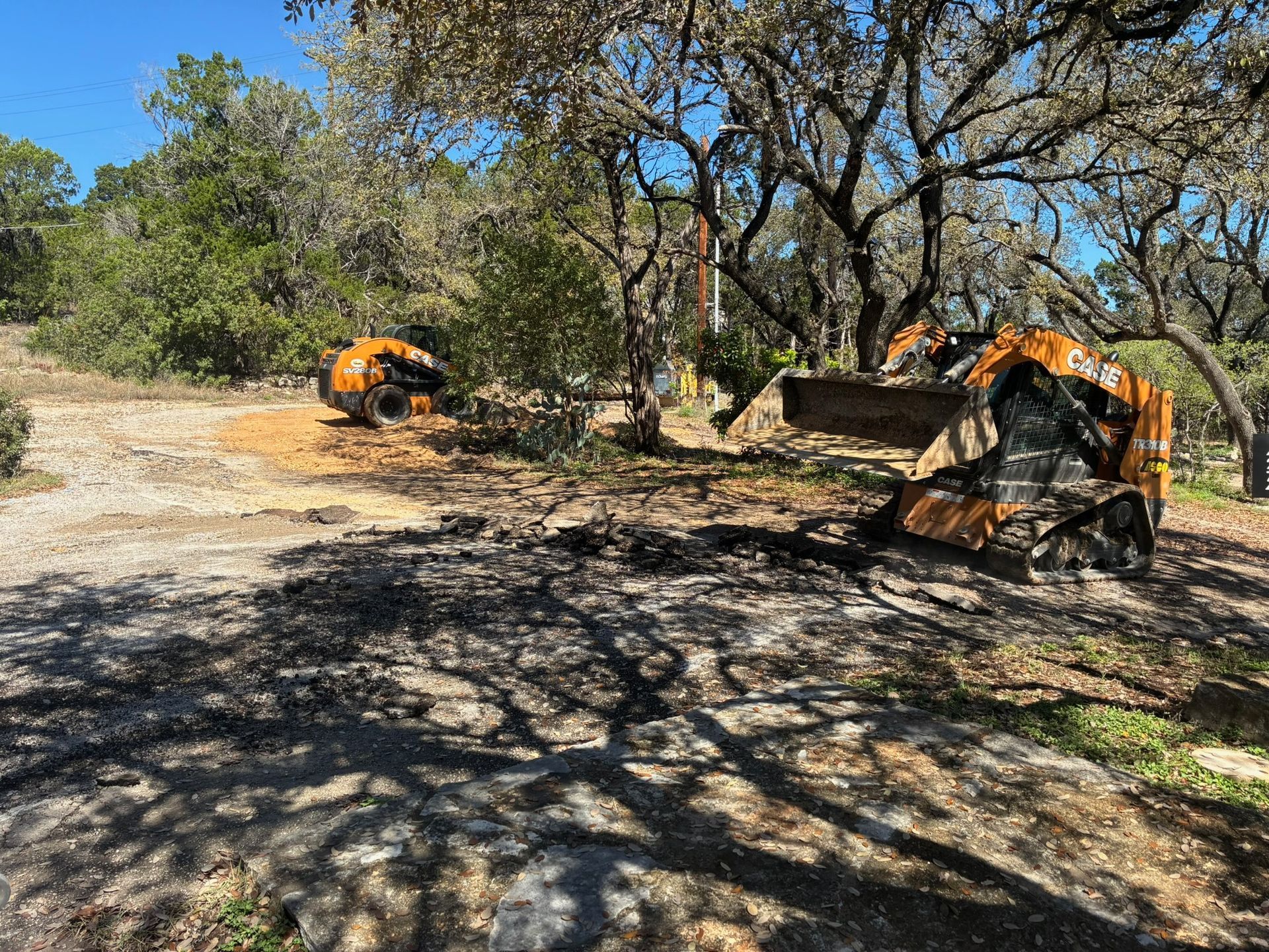 Two orange skid-steer loaders clear brush and dirt in a wooded, sunlit lot.