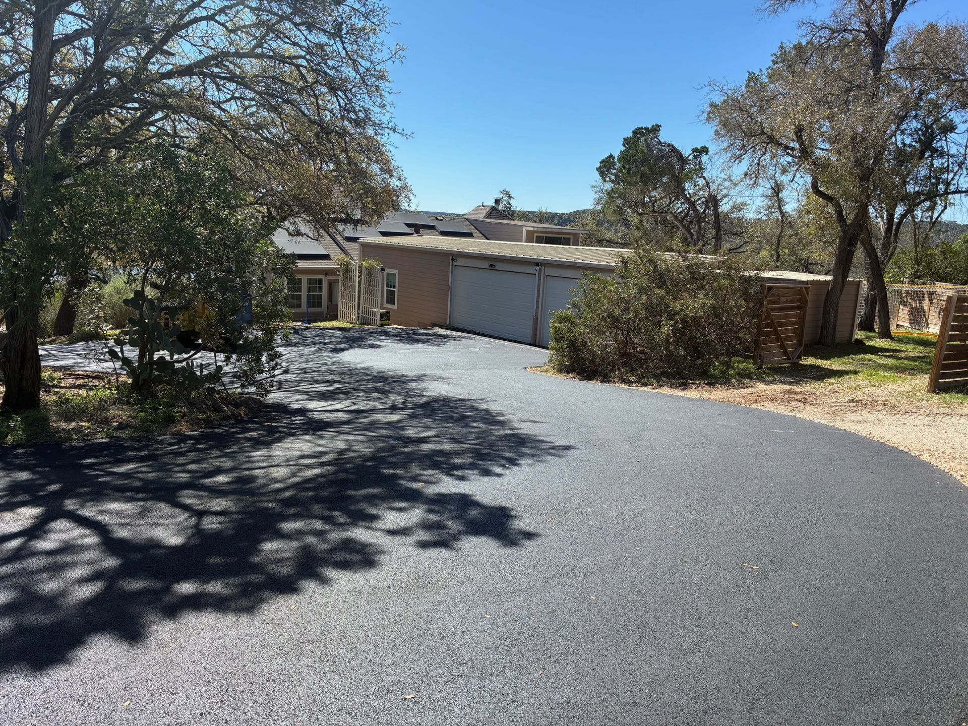 A freshly paved black asphalt driveway leads toward a tan house with a multi-car garage on a sunny, tree-lined property.
